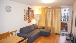 Een moderne woonkamer met een grijze hoekbank, een houten eettafel met stoelen, een wandklok en een groot raam met gordijnen.