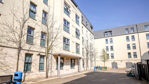 A courtyard with a building featuring multiple windows and a flat roof. There are trees, a bench, and a trash bin visible. - Popup Bath (Bath)