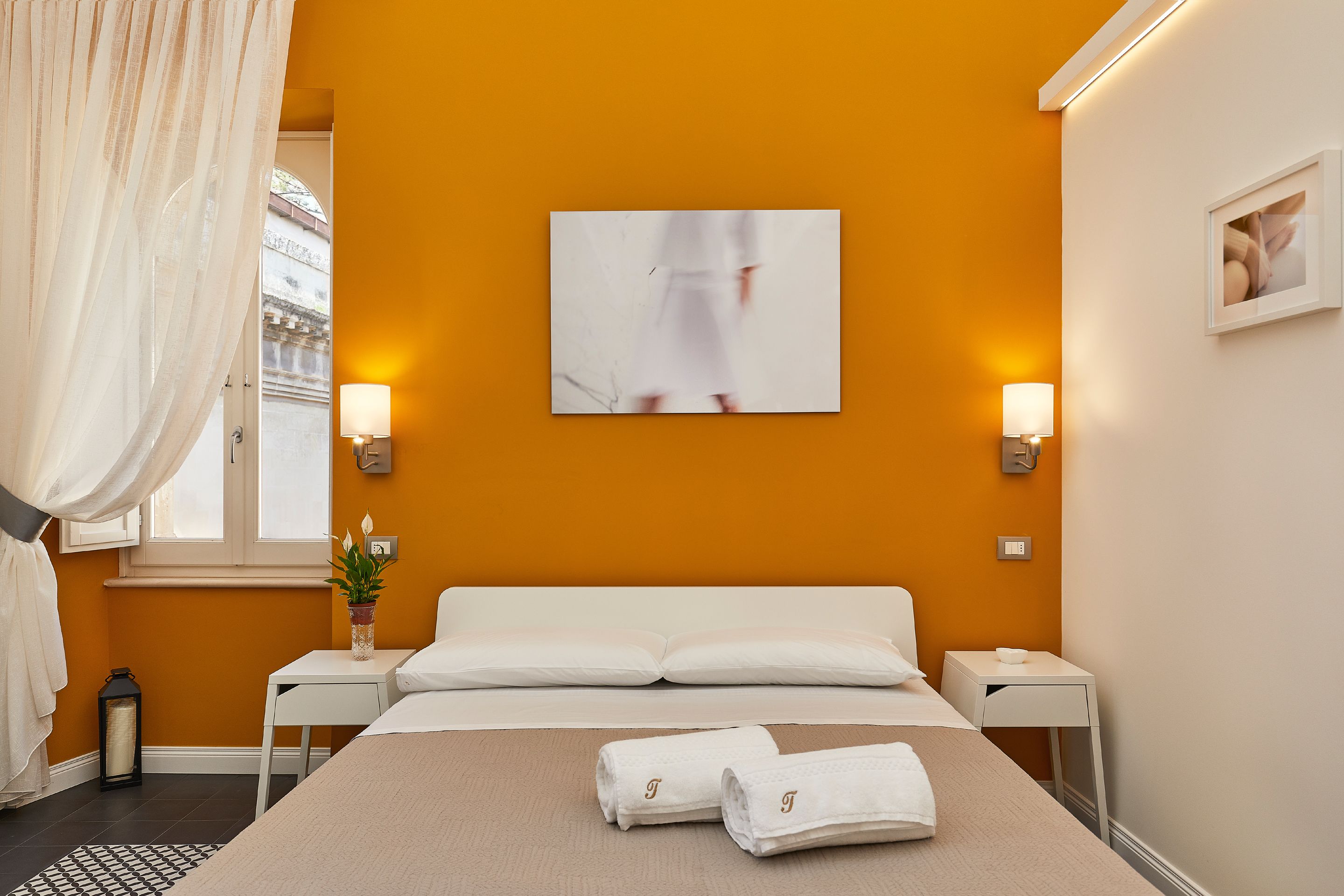 A hotel room with a bed, two bedside tables, a window with curtains, a wall-mounted picture, and two wall sconces.