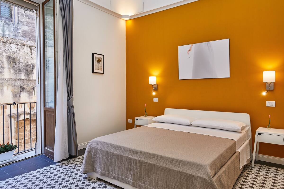 Ein Schlafzimmer mit einem Bett, zwei Nachttischen, einem Fenster mit Vorhängen und einem an der Wand hängenden Bild.