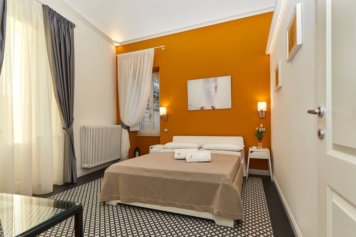 Ein Hotelzimmer mit Bett, Nachttischen, Heizkörper, einem Fenster mit Vorhängen und einer Wandleuchte.