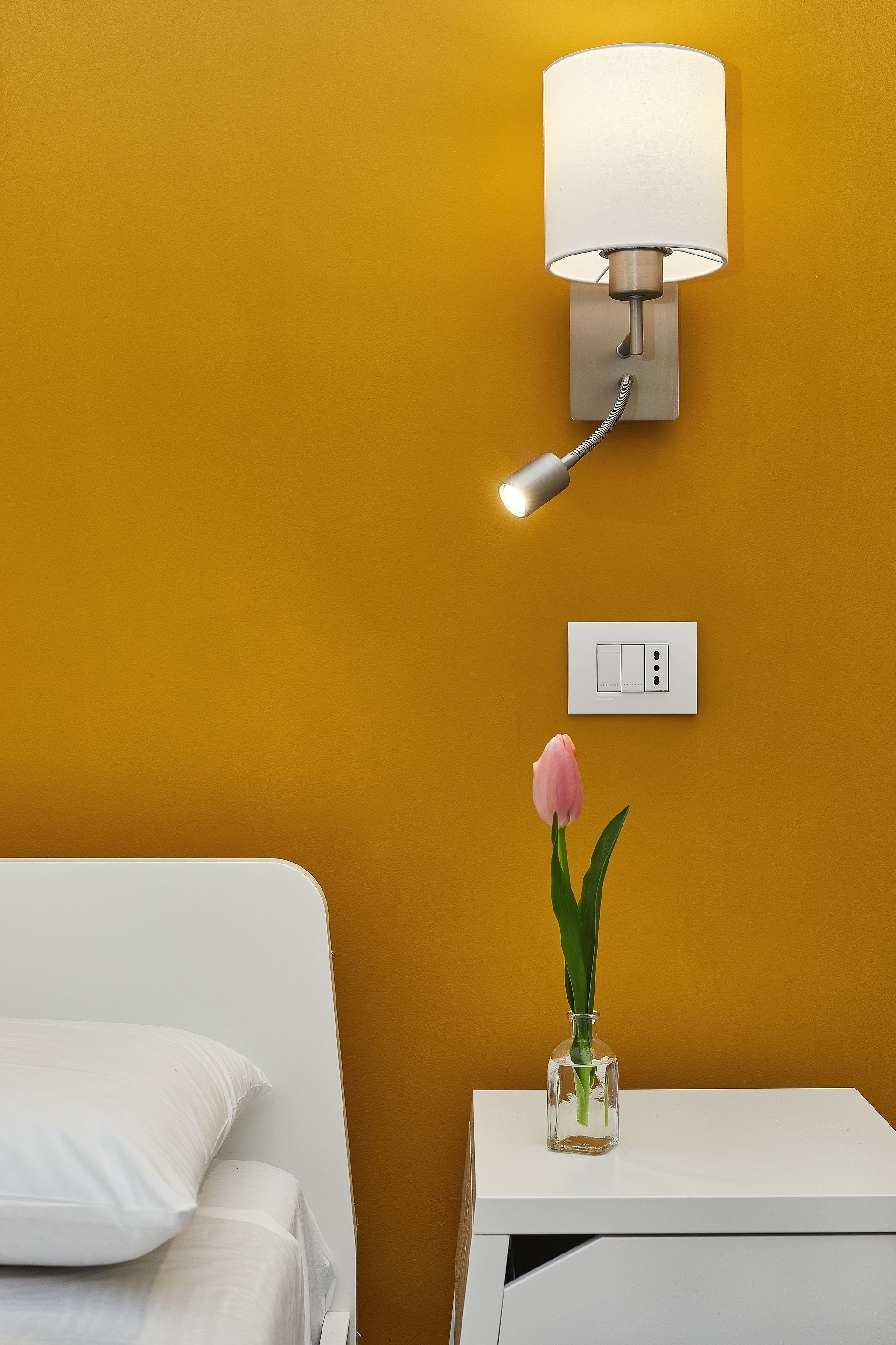 Habitación de hotel con cama, mesita de noche, lámpara de pared, toma de corriente y un jarrón con una flor.