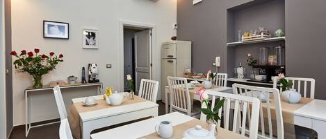 A dining area with white tables and chairs, a refrigerator, and a shelf with kitchen items.