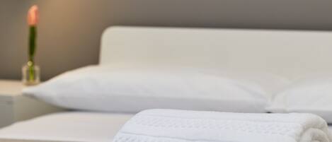 A neatly made bed with white linens and a single pillow, a bedside table with a lamp and a vase of flowers, and a wall-mounted artwork.