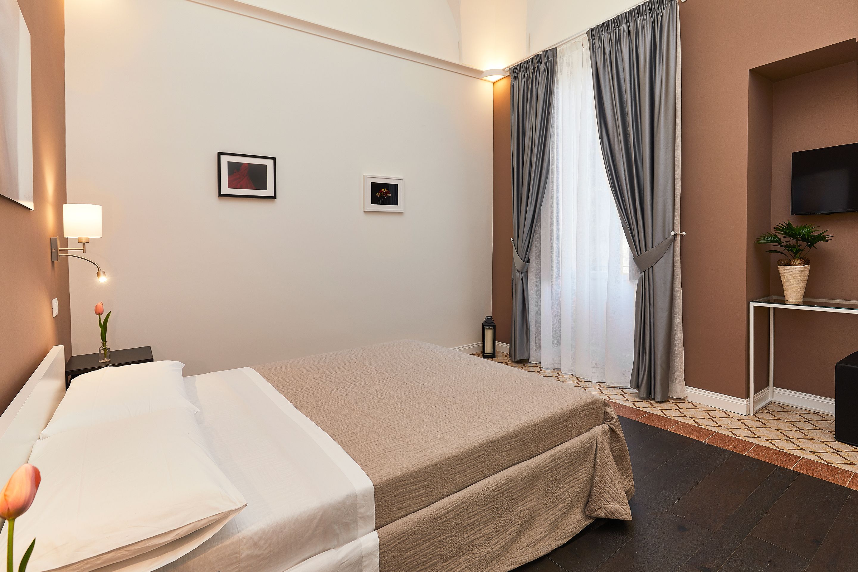 A hotel room with a bed, a nightstand with a lamp, a small table with a plant, and a TV mounted on the wall.