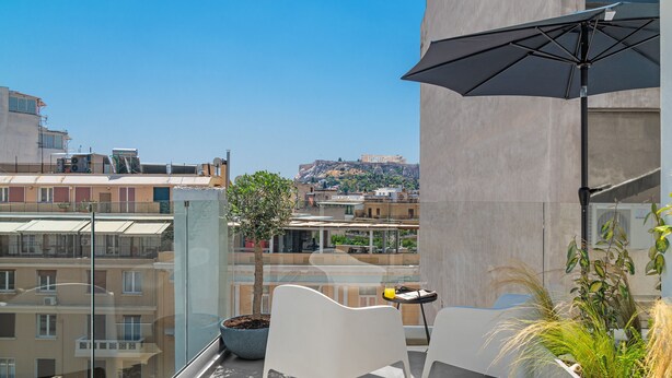 Un balcón con un árbol en maceta, dos sillas blancas, una mesa con un objeto amarillo y vistas de un edificio histórico.