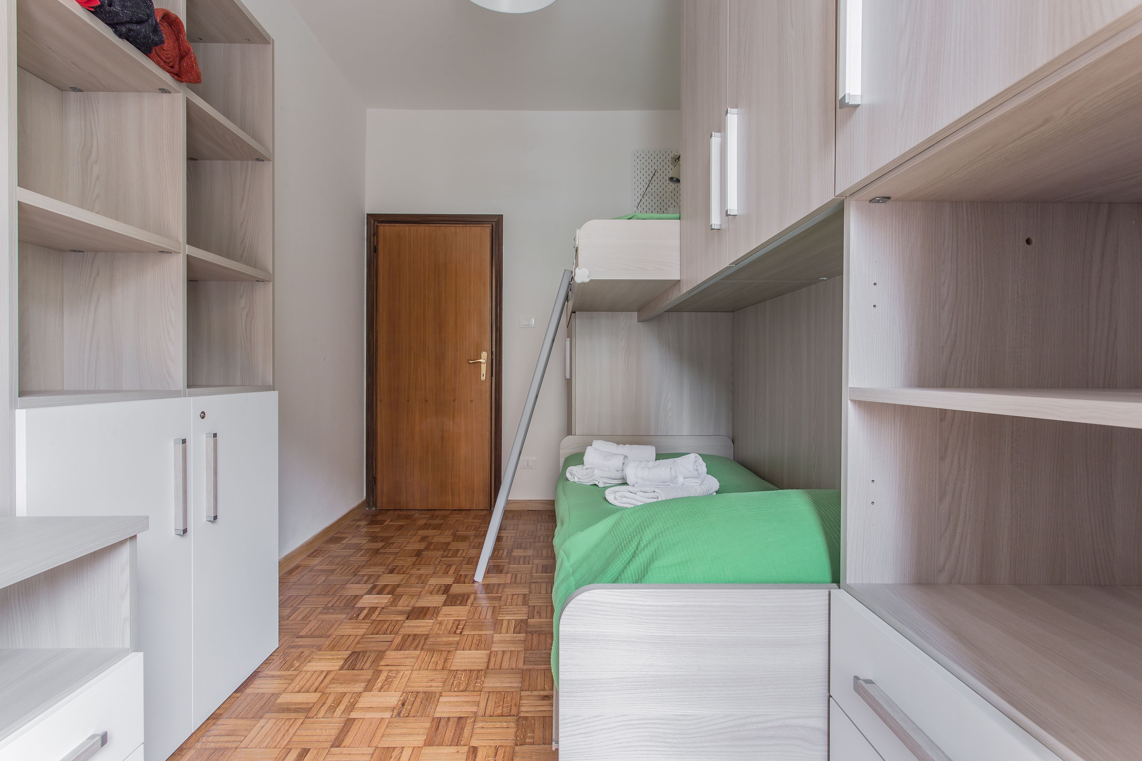 A narrow room with a bunk bed, wooden flooring, and built-in shelving.