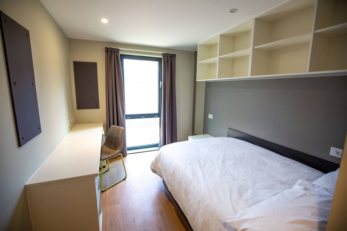 A hotel room with a bed, a desk, a chair, and a large window with curtains.