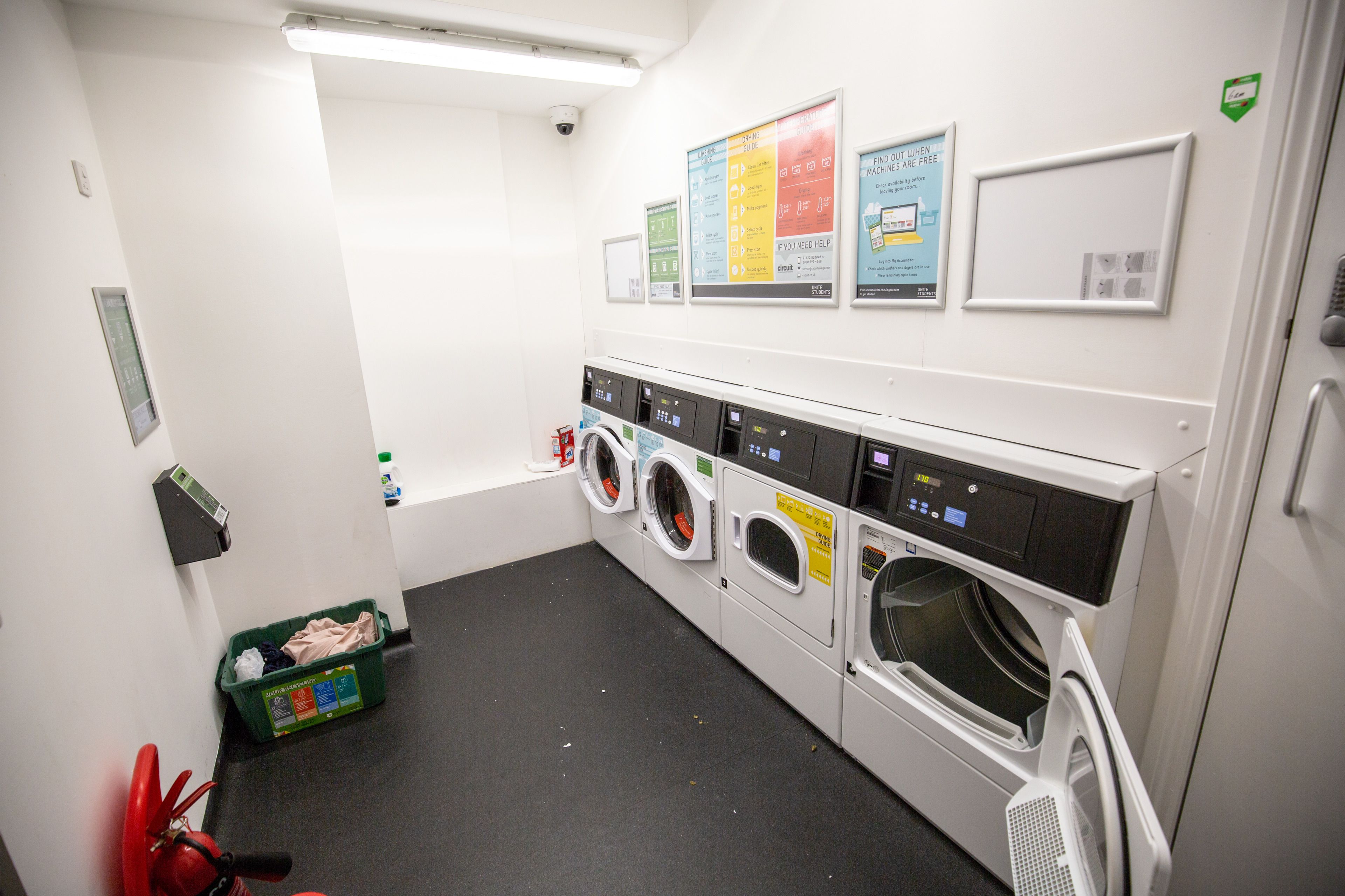 A laundry room with multiple washing machines, a fire extinguisher, a green bin, and informational posters on the wall.