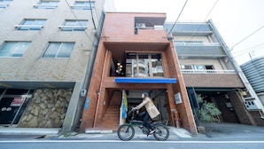 A brick building with a blue awning, a sign indicating a hostel, and a person riding a bicycle. - HOSTEL PAQ tokushima - Hostel (Tokushima)