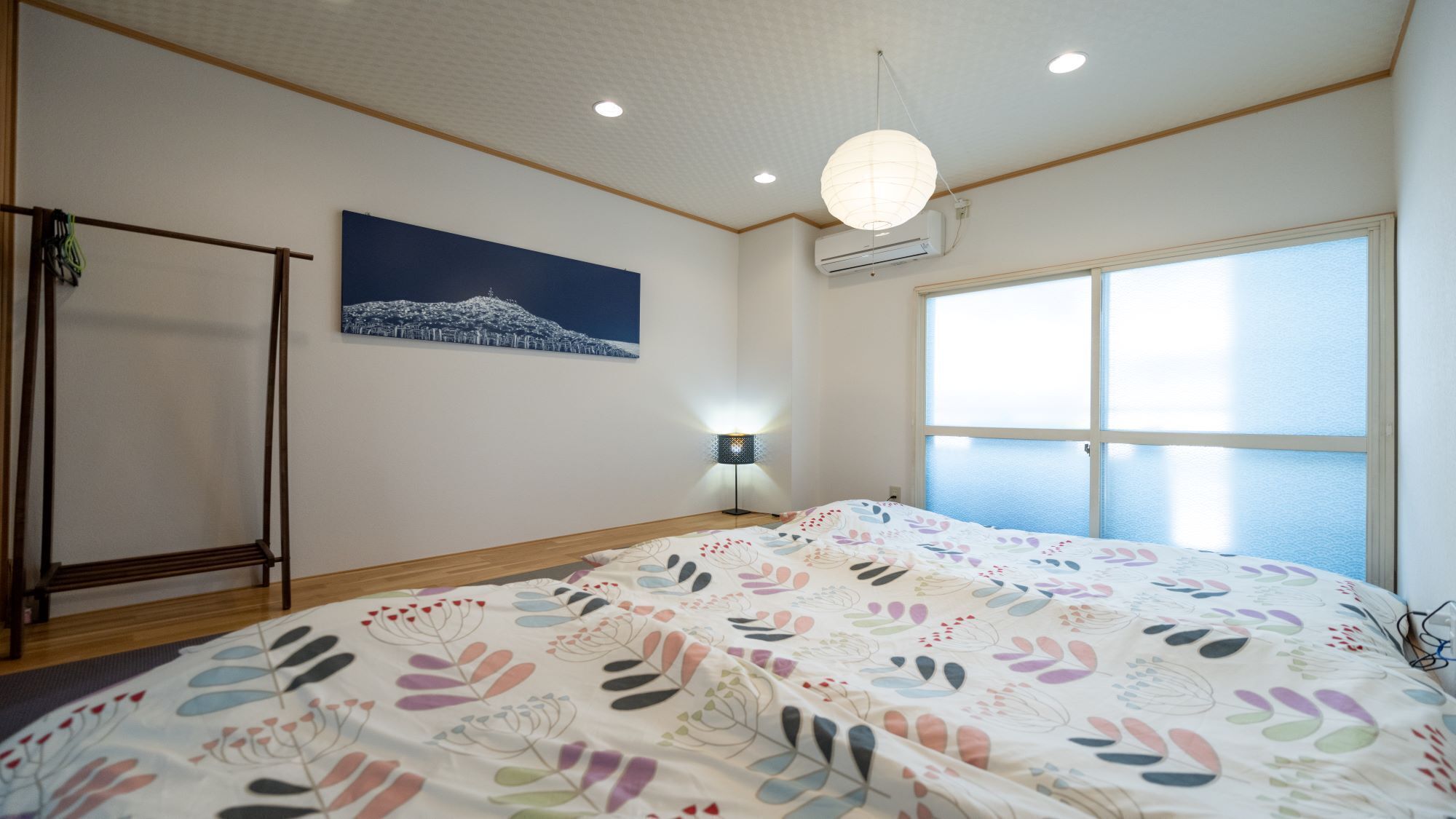 Un dormitorio con una cama, una mesita de noche, una lámpara colgante, una ventana con vistas y un cuadro en la pared.