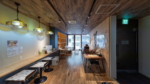 A modern cafe with wooden floors and walls, featuring tables and chairs, hanging lights, and a view of the city through large windows. - HOSTEL PAQ tokushima - Hostel (Tokushima)