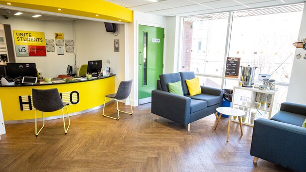 A reception area with a bright yellow wall, a green door labeled "First Floor," and a seating area with blue sofas and a small round table. - Popup Liverpool (Liverpool)
