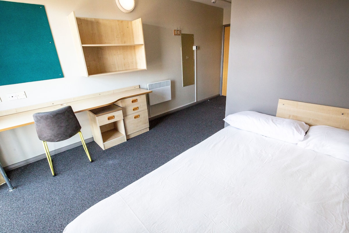 A hotel room with a bed, a desk with drawers, a chair, a mirror, and a door.
