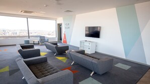 Un espace de bureau moderne avec des canapés gris, un téléviseur à écran plat et une vue sur la ville grâce à de grandes fenêtres.