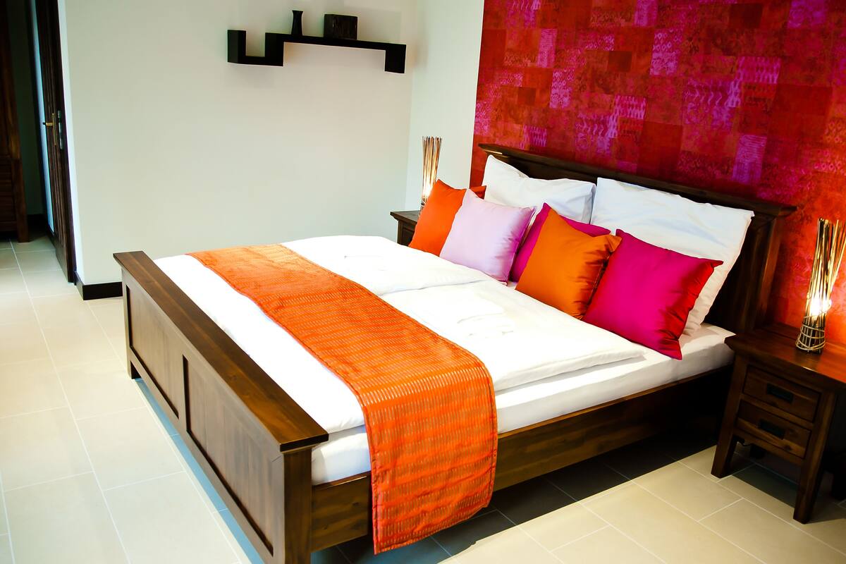 Ein ordentlich bezogenes Bett mit einer orangefarbenen Tagesdecke, einem Holzkopfende und mehreren Kissen in verschiedenen Farben.