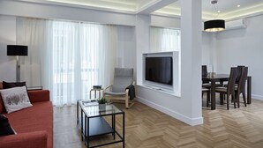 A modern living room with a sofa, a glass coffee table, a TV, a dining area with chairs, and a large window with curtains. - Luxurious Spacious Apt next  Hilton Area (Athens)