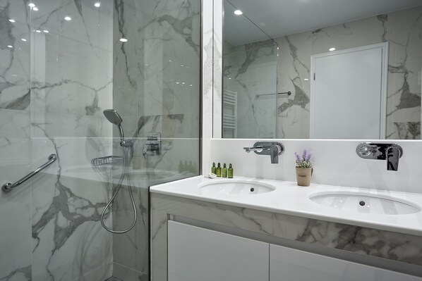 Un baño moderno con paredes de mármol, un espejo grande, dos lavamanos y una ducha.