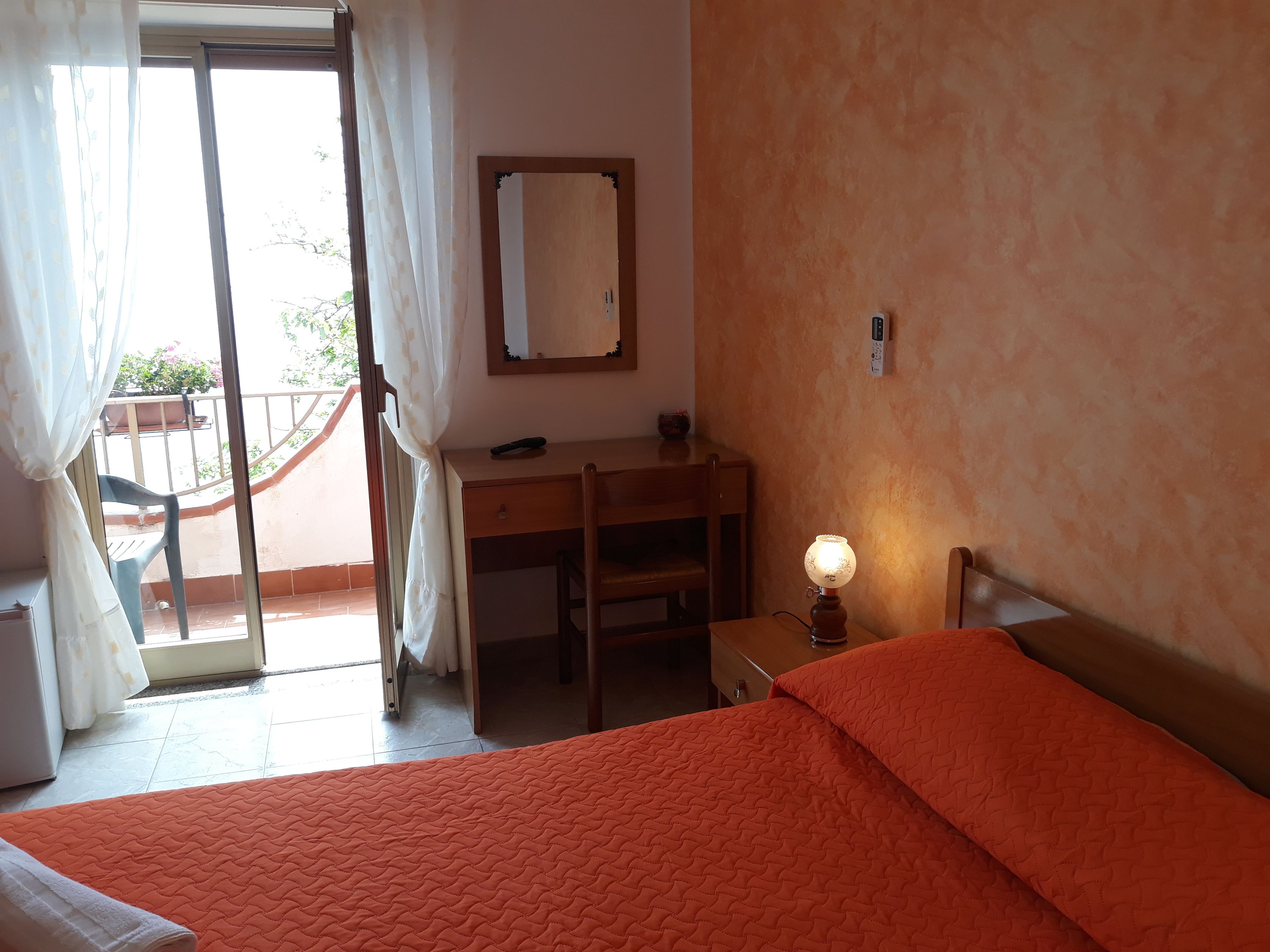 A bedroom with a bed, a desk, a chair, a nightstand, a lamp, and a window with curtains.