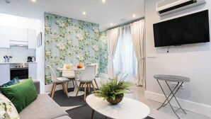 A modern living room with a sofa, a dining table with chairs, a small round table with a plant, a TV mounted on the wall, and an air conditioning unit. - Feelathome San Marcos Apartments (Madrid)