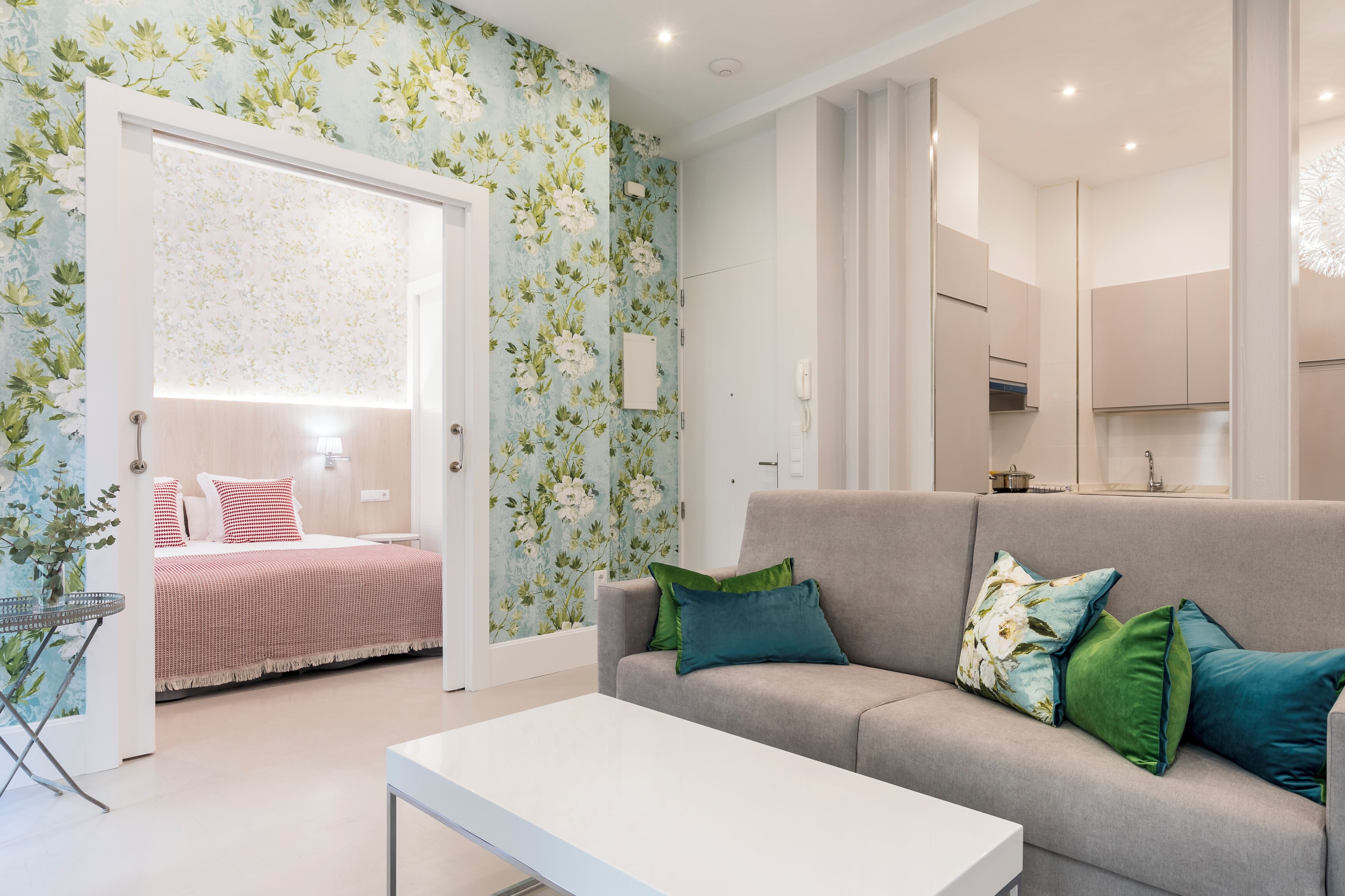 A modern living room with a sofa, green and blue pillows, a white coffee table, and a bed with a red and white checkered blanket.
