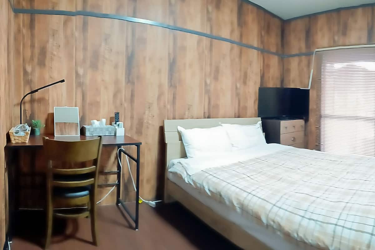 Quarto com cama, escrivaninha com cadeira, televisão e janela com persianas.