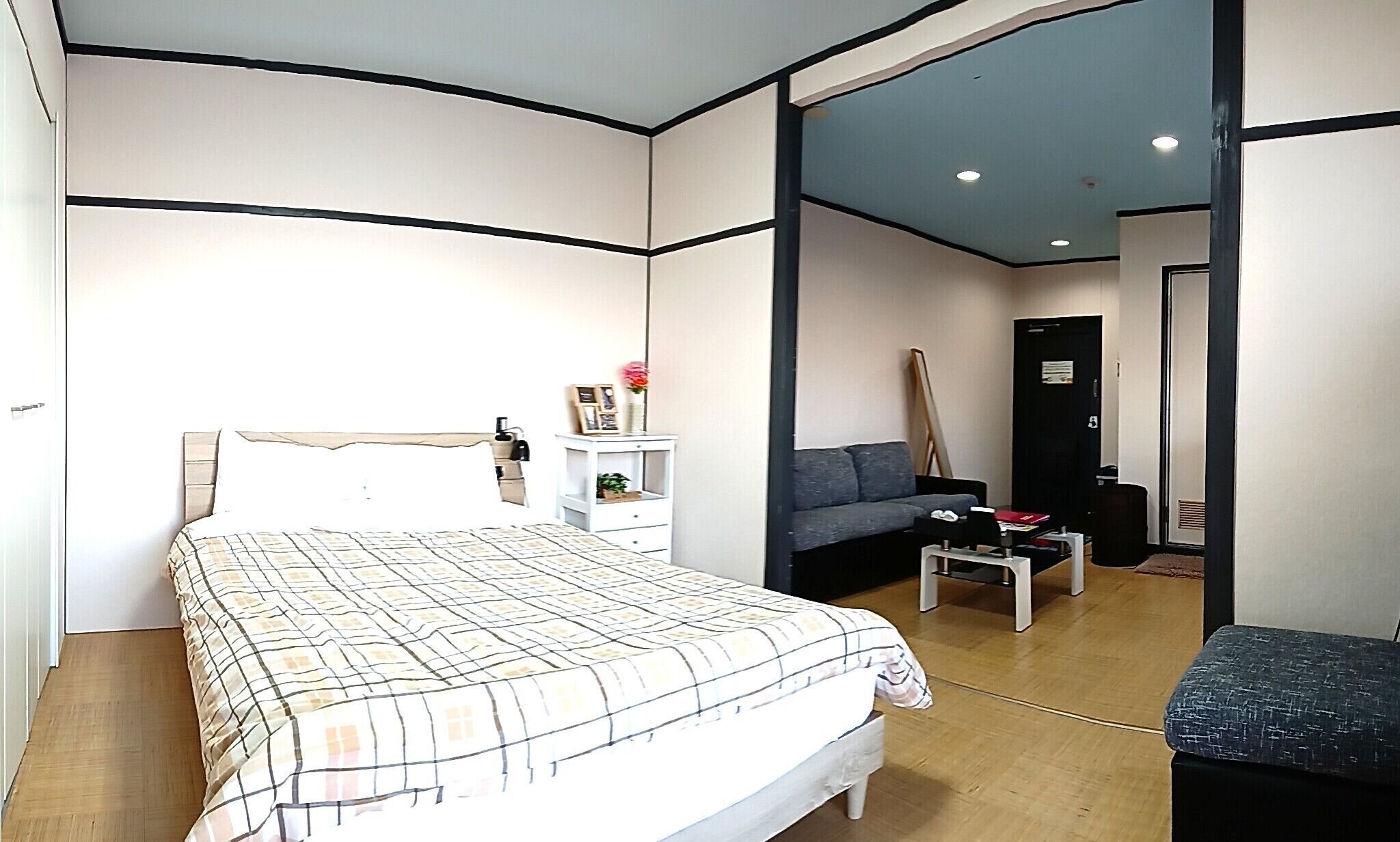 A bedroom with a bed, a nightstand, a dresser, a gray sofa, a small table, and a door leading to another room.