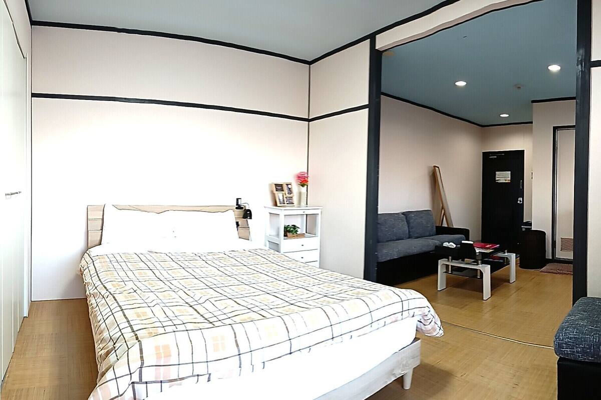 Quarto com cama, mesa de cabeceira, cômoda, sofá cinza, pequena mesa e porta que dá para outro cômodo.