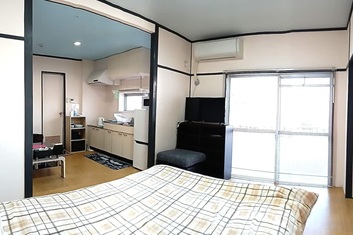 Quarto de hotel compacto com cama, pequena mesa, cozinha compacta, televisão e janela com persianas.
