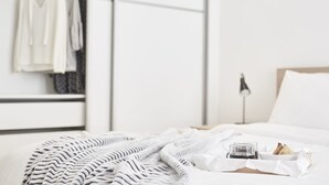 Ein ordentlich bezogenes Bett mit einer gestreiften Decke, ein Tablett mit Toast und einem Glas Wasser sowie ein Kleiderständer in einem minimalistisch eingerichteten Raum.