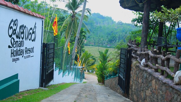 A pathway leading to a gated entrance of a resort named "Galapagos Senoro Holiday Resort" with a lush green landscape in the background. - Senaro Holiday Resort (Udugama)