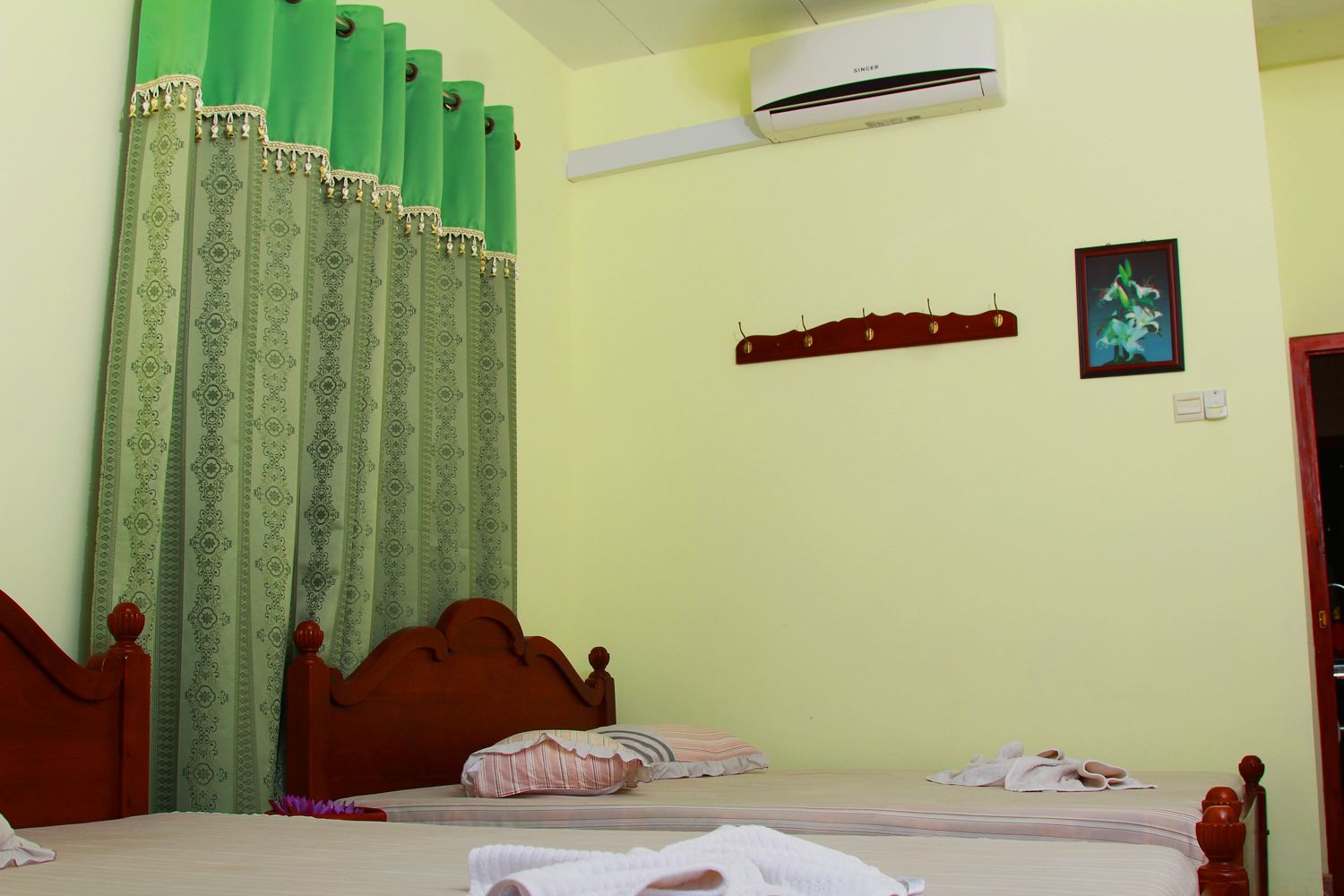 A bedroom with a bed, a green curtain, an air conditioning unit, a wooden headboard, a bedside table, and a wall-mounted shelf with a picture.