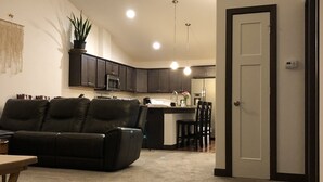 Una sala de estar moderna con un sofá de cuero oscuro, una mesa de comedor y una zona de cocina con armarios oscuros y una encimera blanca.