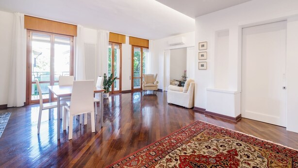 A spacious living room with a wooden floor, a dining area with white furniture, and a large window with curtains. - Padova - Via Faggin 47A (Padova)