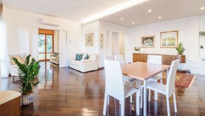 A spacious living area with a wooden dining table, white chairs, and a sofa. There are potted plants and framed pictures on the walls. - Padova - Via Faggin 47A (Padova)