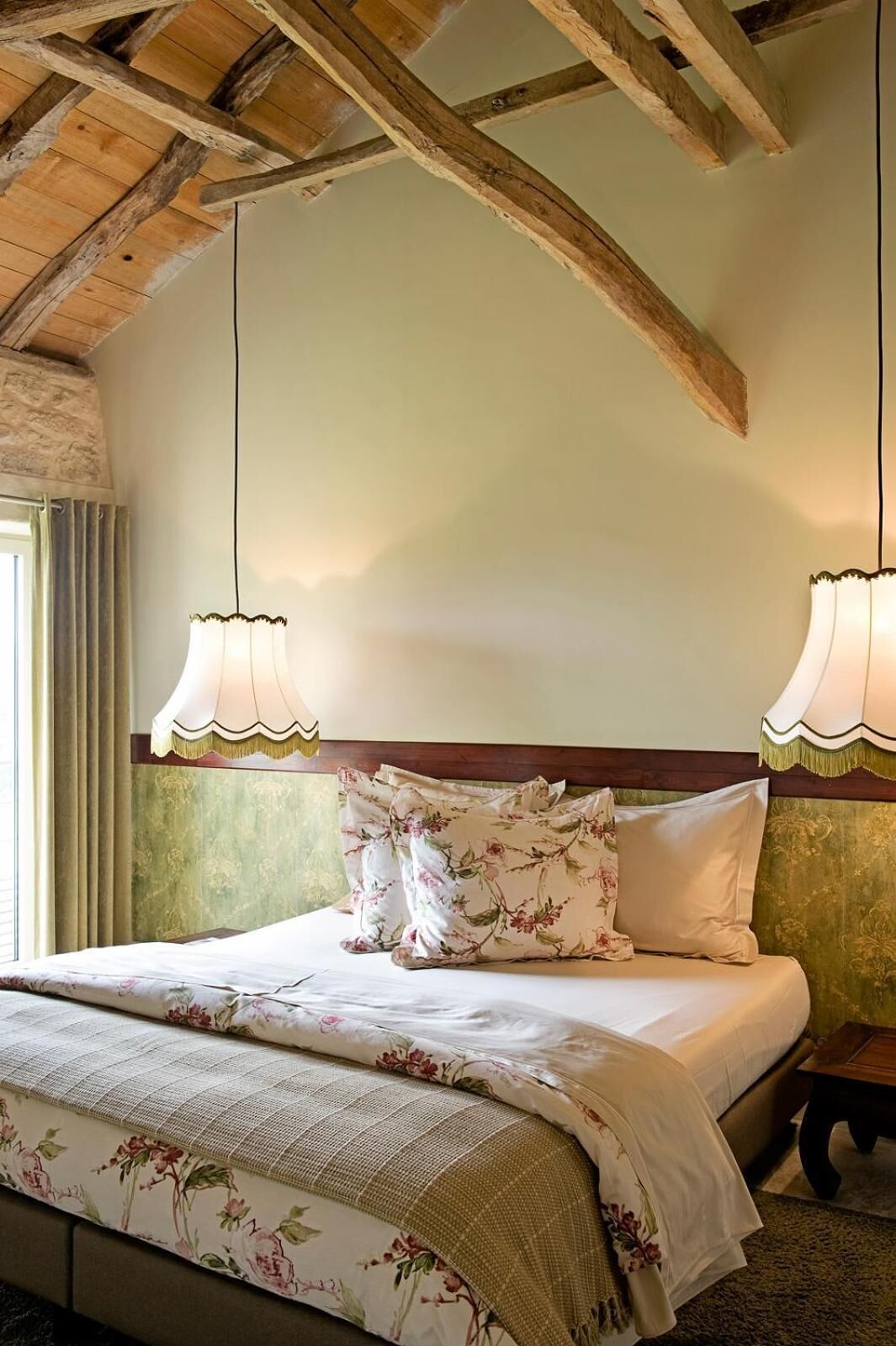Un dormitorio con techo de madera, una cama con ropa de cama floral, dos lámparas de pared y un ventanal con cortinas.