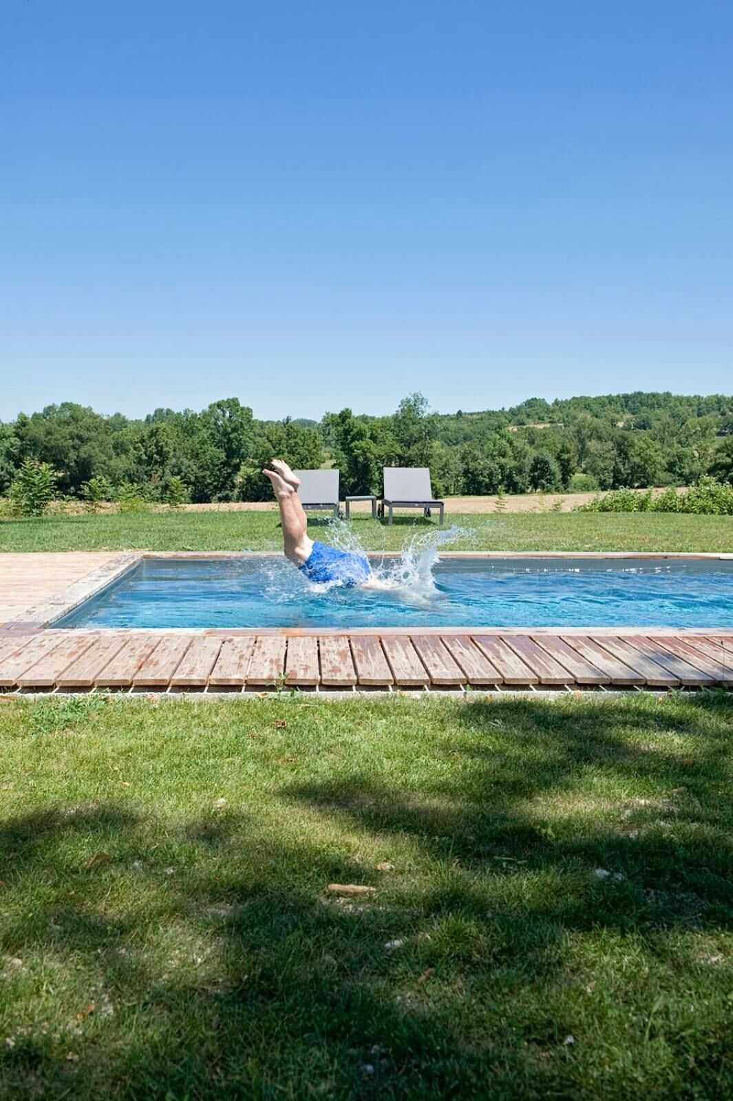 Una piscina con muelle de madera, rodeada de césped y árboles.