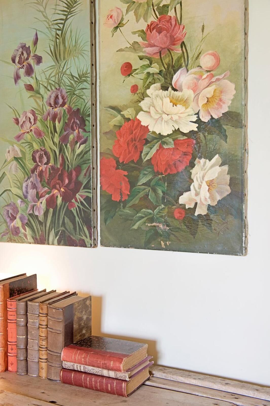 A wall with two floral paintings and a stack of old books on a wooden surface.