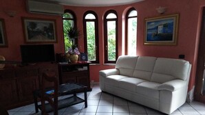 A living room with a white sofa, a wooden table, a TV, and a window with a view of greenery. - Bed & Breakfast Monti e Mare (Lanusei)