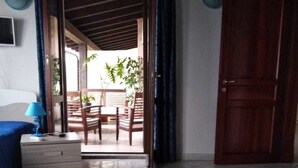 A room with two beds, a desk with a lamp, a wooden door, and an open door leading to an outdoor area with chairs and plants. - Bed & Breakfast Monti e Mare (Lanusei)