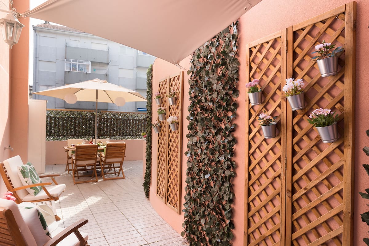 A patio area with wooden furniture, a table, and chairs. There are potted plants on a lattice wall.