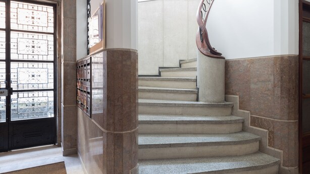 Una imponente escalera de mármol con barandilla de madera, que conduce a una plataforma con un amplio ventanal y una puerta con celosía de metal.