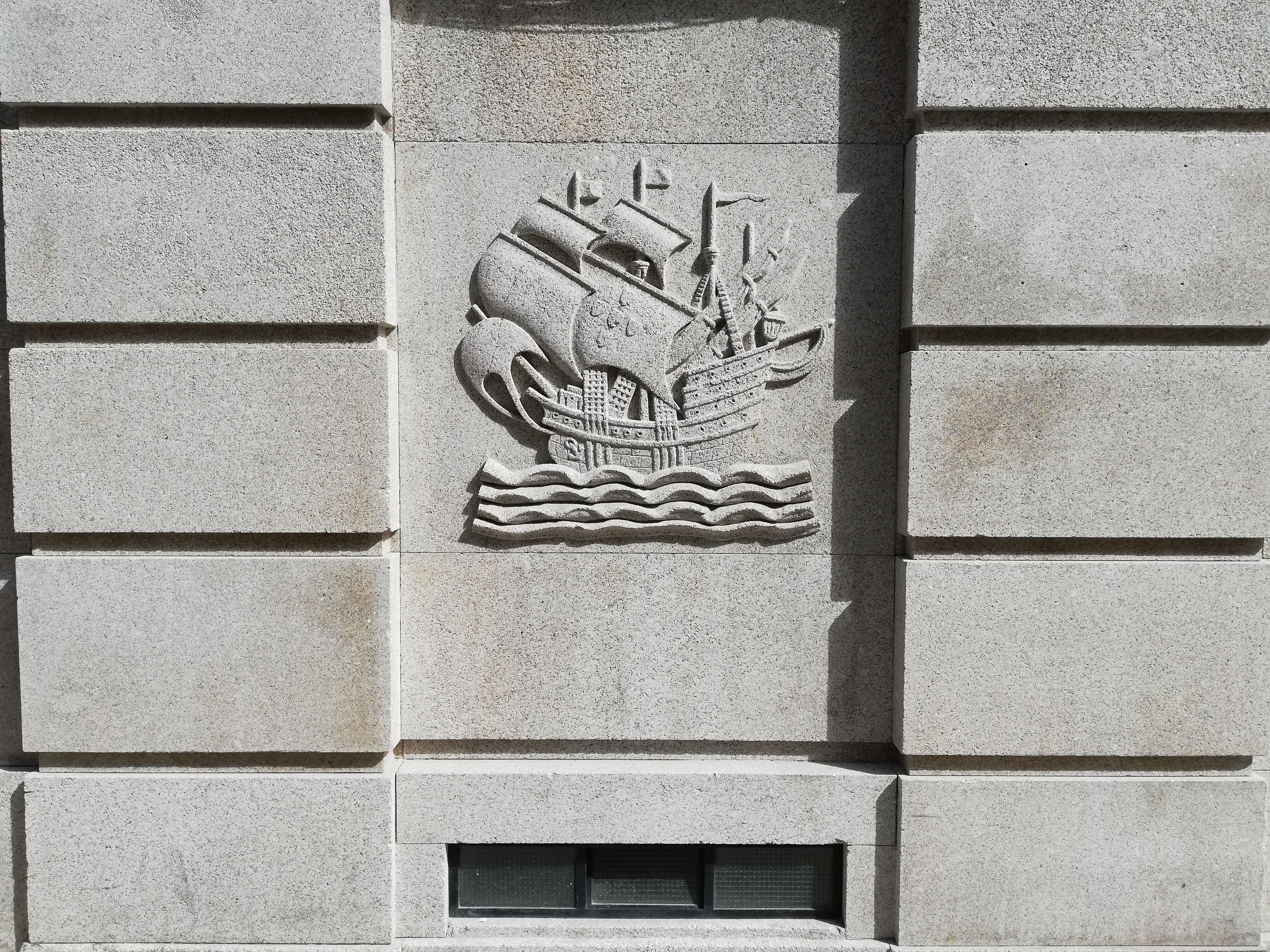 Un bas-relief en pierre représentant une main tenant un navire, ornant la façade d’un bâtiment.