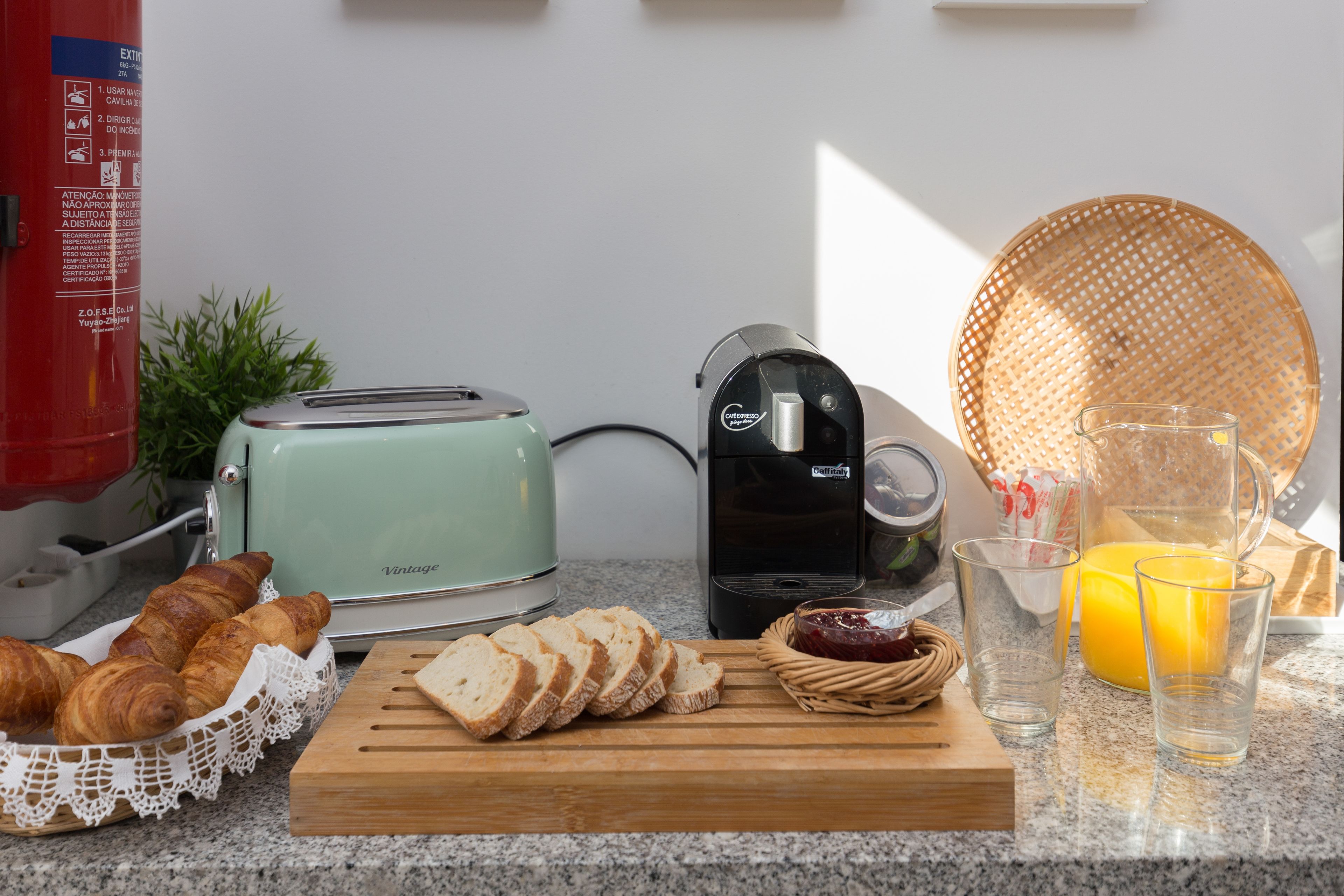 A kitchen counter with a toaster, coffee maker, croissants, sliced bread, a cutting board, a wicker basket, a glass of orange juice, and a glass of water.