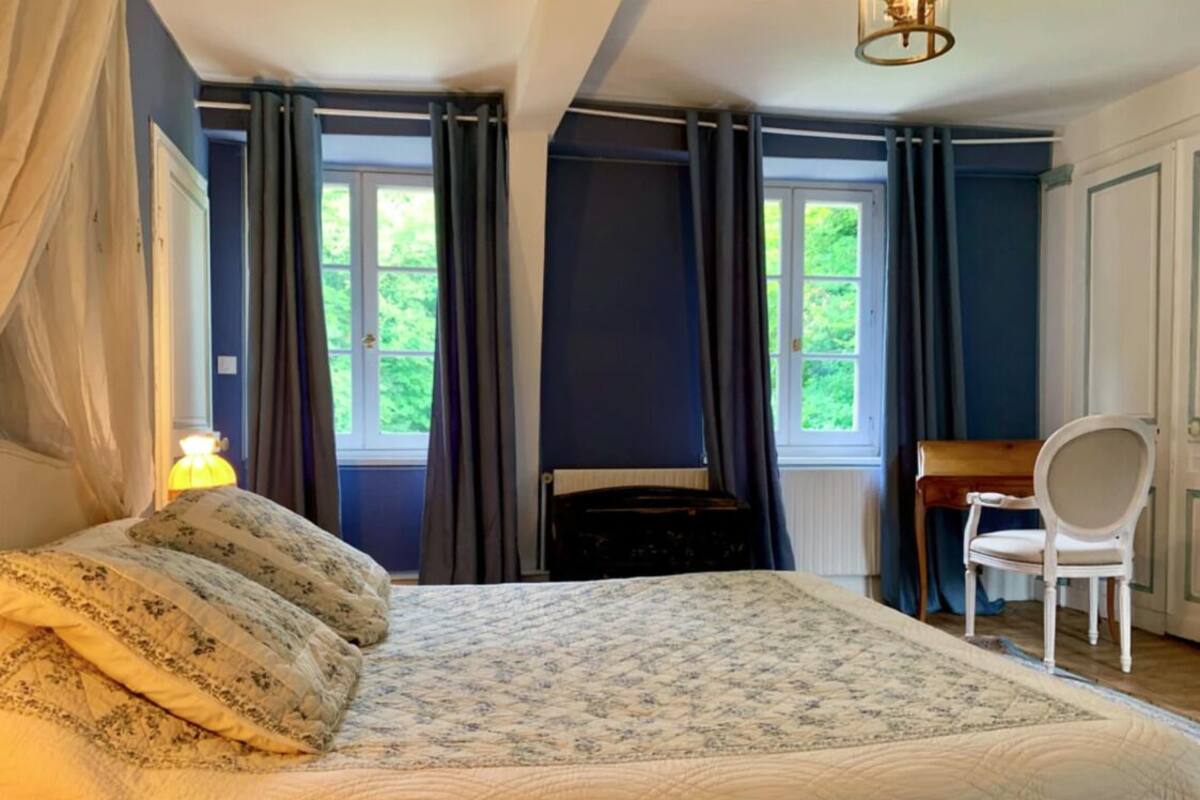 Une chambre à coucher avec un lit, une chaise, un bureau et deux fenêtres avec des rideaux.