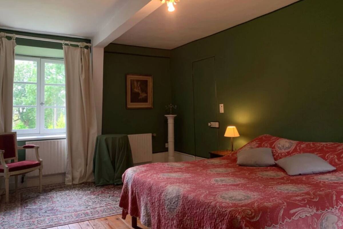 Une chambre à coucher avec un lit, une chaise, une fenêtre avec des rideaux, une lampe et un tableau au mur.