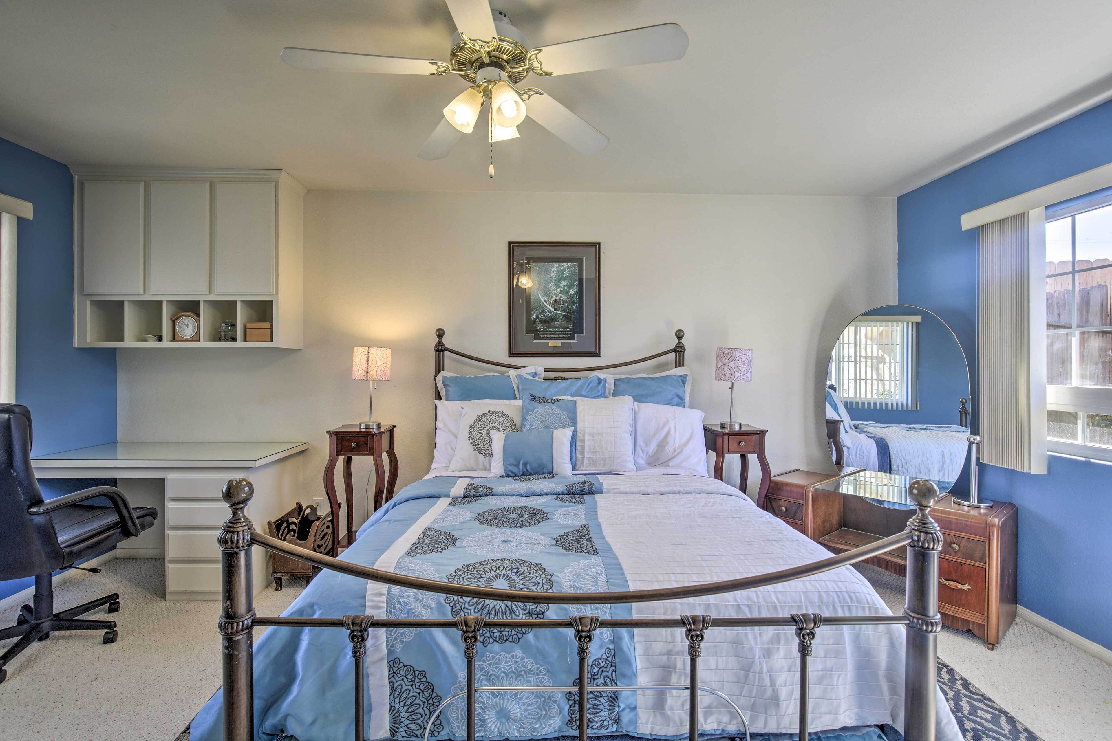 A bedroom with a bed, a desk, a chair, a ceiling fan, a framed picture, and a window with a view.