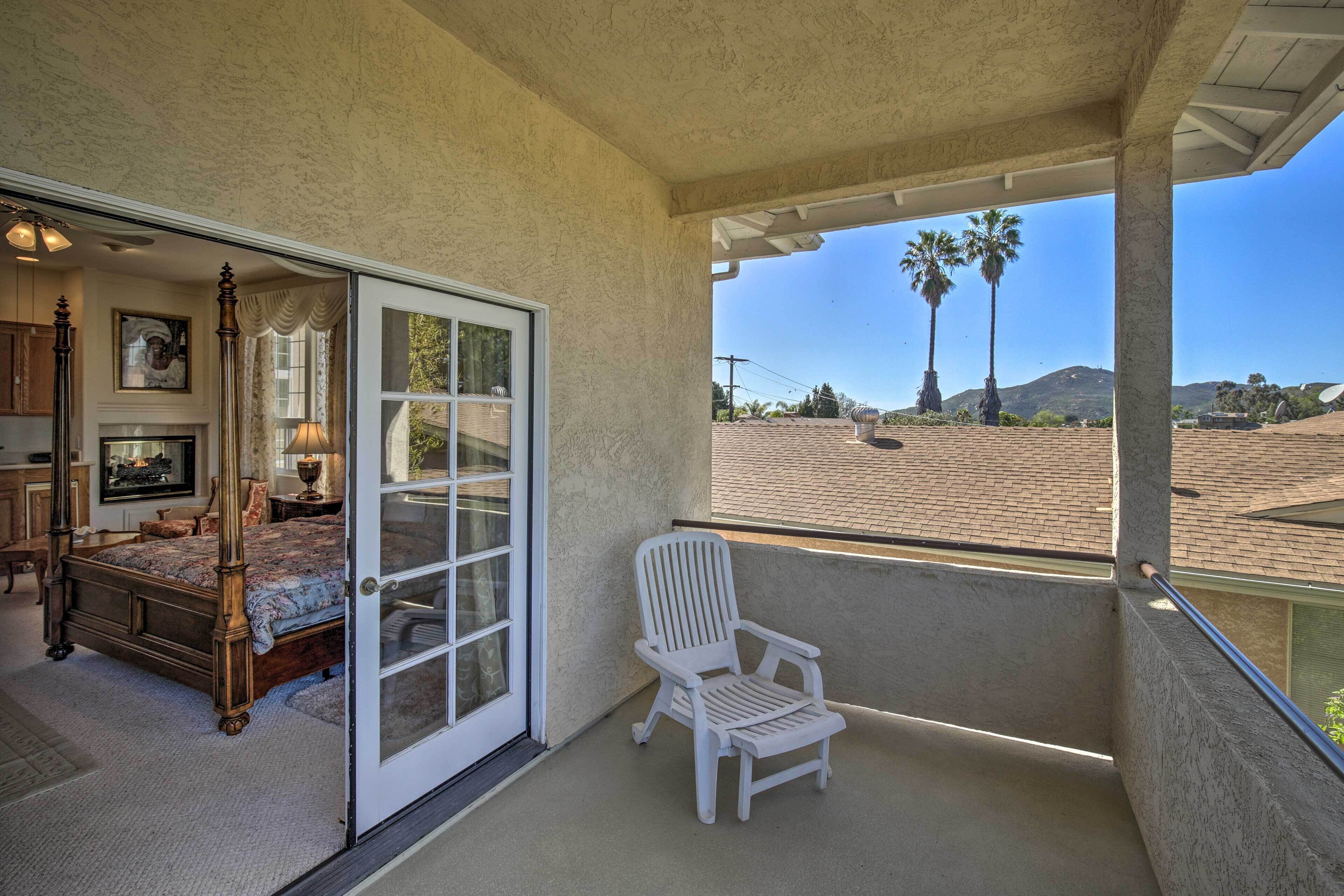 A bedroom with a bed, a chair, and a sliding glass door leading to a balcony with a view of palm trees and a mountain.