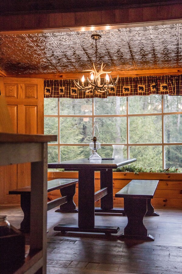 Ein rustikales Interieur mit Holzbänken und einem Tisch, einer Kristallleuchtre und einem großen Fenster mit Blick auf Bäume.