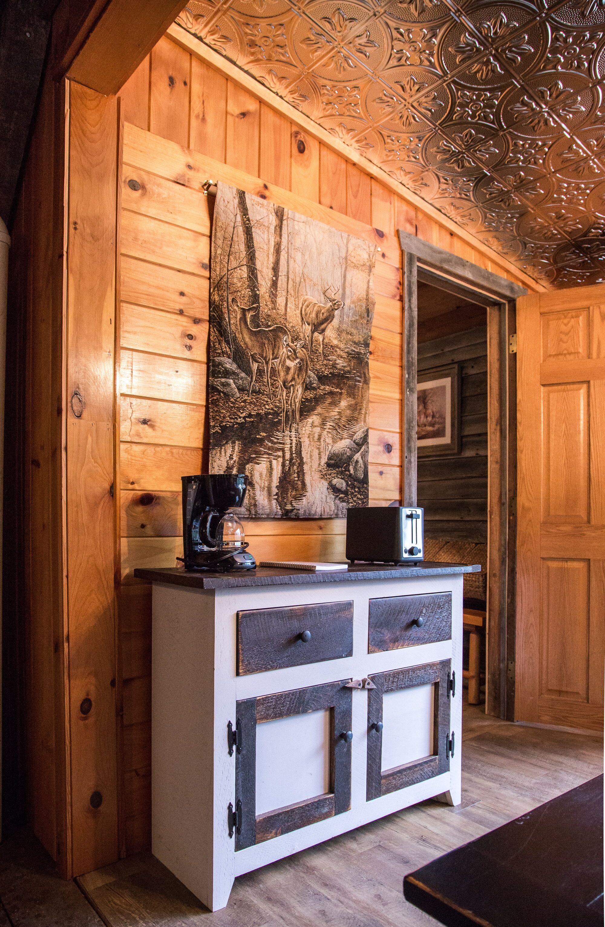 Interior de una cabaña de madera con pared rústica, techo decorativo de metal, un armario de madera con cajones y un cuadro de animales salvajes colgado encima.